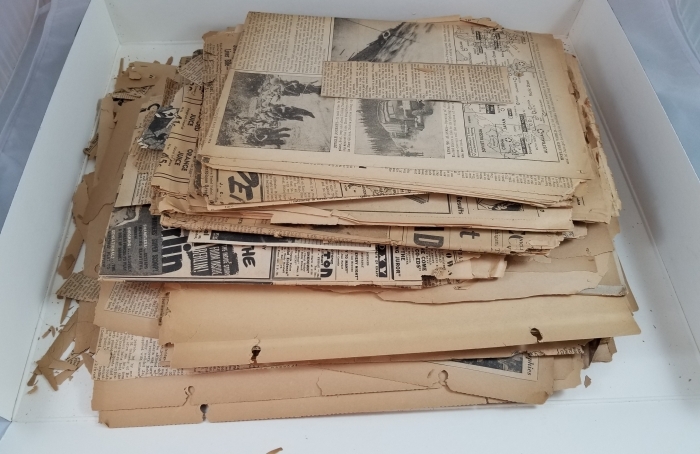 A box contains a stack of yellowed and crumbling newspapers and scrapbook pages.