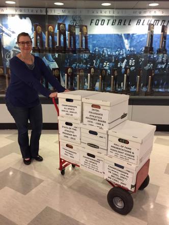 An archivist standing with a cart of record boxes in front of a trophy case